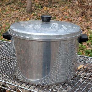 Può includere: Una grande pentola da cucina color argento con coperchio e manici neri. La pentola è appoggiata su una griglia metallica, con uno sfondo naturale all'aperto.