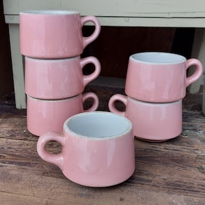 Könnte beinhalten: Sechs rosafarbene Keramikbecher mit weißem Innenraum. Die Becher sind pyramidenförmig gestapelt, wobei der oberste Becher der kleinste und der unterste Becher der größte ist. Die Becher sind alle gleich groß und gleich geformt und haben ein schlichtes, klassisches Design.