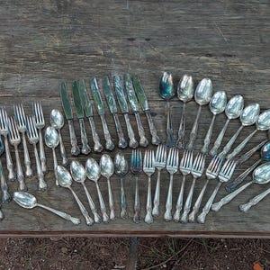 May include: An assortment of antique silverware, including forks, knives, and spoons, arranged on a weathered wooden surface. The silverware has ornate handles and a silver finish, reflecting light. A collection of vintage cutlery.