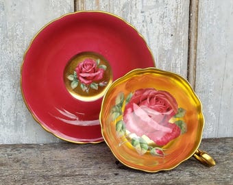 May include: A red teacup and saucer with gold trim and a floral design featuring a red rose. The saucer has a gold rim and a red centre with a smaller gold rim and a single red rose in the centre.