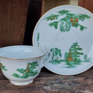 Può includere: Una tazzina da tè e un piattino bianchi con un disegno verde e oro di una pagoda e alberi. Il disegno ricorda un giardino giapponese tradizionale.