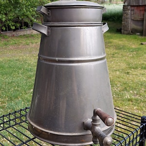 May include: A vintage silver-colored metal water cooler with a wooden handle on the spigot. The cooler has a double-tiered lid and sits on a black metal stand.