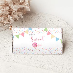Może przedstawiać: Prostokątne opakowanie na batonik z białym tłem, kolorową posypką, pastelowymi chorągiewkami oraz napisem "SHE'S A Sweet one", gdzie mały różowy pączek zastępuje literę "o" w słowie "one".