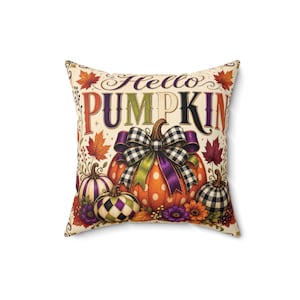 Peut inclure: Un coussin carré décoré avec le texte Hello PUMPKIN en lettres stylisées. Le motif présente une grande citrouille orange à pois avec un nœud à carreaux noir et blanc, entourée de citrouilles plus petites, de feuilles d'automne et de fleurs.