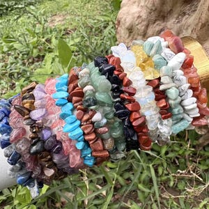 May include: A collection of gemstone chip bracelets in various colors, including blue, purple, red, green, and white. The bracelets are displayed on a white cylindrical object against a backdrop of green foliage.