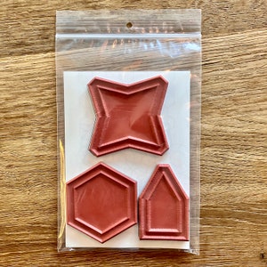Peut inclure: Trois tampons de formes géométriques dans un sac en plastique transparent. Les tampons sont de couleur brun rougeâtre et comprennent une étoile, un hexagone et une forme de maison. Les tampons sont sur un fond blanc avec un motif subtil.