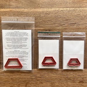 Op de afbeelding: Drie doorzichtige plastic zakken met rode rubberen stempels met een geometrisch ontwerp. Elke stempel staat voor een witte kaart. De zakken liggen op een houten oppervlak. De stempels zijn voor VersaCraft-stempelen.