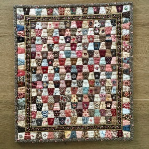 May include: A handcrafted patchwork quilt with a grid pattern of small, colourful fabric pieces. The quilt features a variety of floral and patterned fabrics in shades of red, pink, brown, and cream. A decorative border frames the quilt.