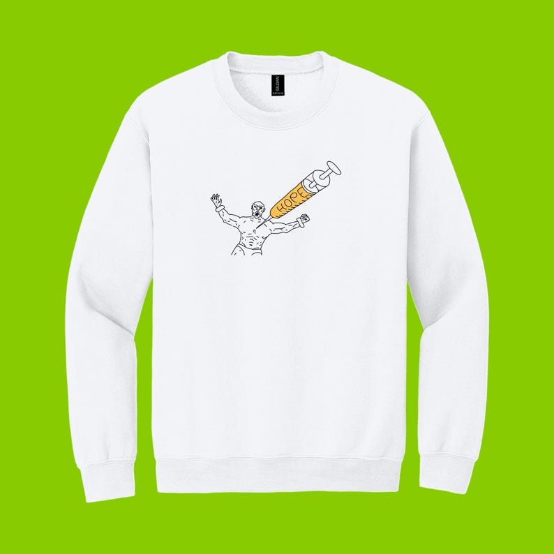 May include: White crewneck sweatshirt featuring a black and orange graphic. The graphic depicts a person with arms raised, with a syringe labeled "HOPE" piercing their chest. The design is a minimalist line drawing style.
