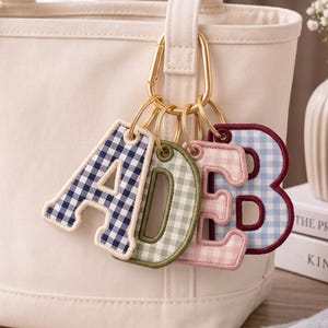 May include: A gold-toned metal carabiner keychain featuring four fabric letter charms in the shapes of A, D, E, and B. Each letter is crafted with a gingham check pattern in navy blue, sage green, light pink, and light blue with stitched borders.