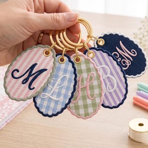 May include: A collection of five oval-shaped fabric keychains with scalloped edges, each featuring a unique embroidered monogram letter. The keychains display various patterns including pink stripes, blue gingham, green gingham, and purple stripes.