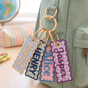 May include: Four rectangular fabric keychains with scalloped edges and gingham patterns in light blue, green, pink, and purple. Each keychain features embroidered names: Sophie, Henry, Alice, and Jasper. They are attached to a gold metal ring.