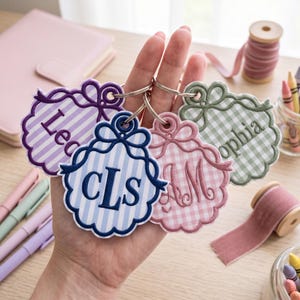 Pode incluir: Quatro porta-chaves de tecido bordado com bordas recortadas e detalhes de laço, apresentando vários padrões e texto personalizado incluindo 'Leo', 'cls', 'AM' e 'ophia'. Cada porta-chaves está preso a um anel de metal prateado.
