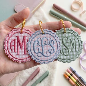 May include: Three circular fabric keychains with scalloped edges and gingham patterns in pink, light blue, and sage green. Each keychain features a centered embroidered monogram in a matching thread color and is attached to a gold metal ring.
