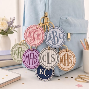 May include: A collection of seven circular, scalloped-edge fabric keychains with embroidered monograms in various colors including pink, blue, green, purple, grey, tan, and navy, all attached to a gold-toned metal ring on a light blue backpack.