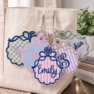 May include: A set of five scalloped, embroidered fabric bag tags attached to a silver metal carabiner. Each tag features a unique pattern, including gingham and stripes, and is personalized with embroidered names like Emily and Sophia.