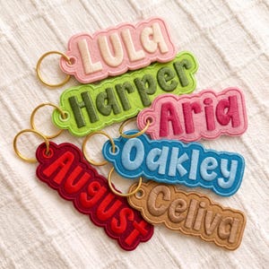 May include: A collection of six embroidered felt keychains with gold-tone metal rings. Each keychain features a scalloped edge and a unique name stitched in a bold, rounded font: LULA in cream, Harper in olive, Aria in pink, Oakley in blue, August in red, and Celiva in tan.