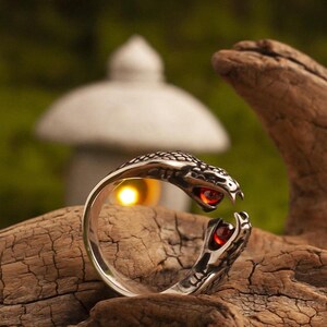 Puede incluir: Un anillo plateado con forma de serpiente, con un patrón de escamas detallado. La cabeza y la cola de la serpiente tienen cada una un ojo de gema roja. El anillo se muestra sobre madera, con un fondo borroso.