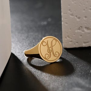 May include: A gold signet ring featuring a circular face with an engraved cursive letter H. The ring has a polished metallic finish and a smooth band that tapers toward the back. The design is centered on the flat circular top surface.