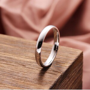 May include: A polished silver ring with a smooth, rounded band. The ring is set against a wooden surface with a soft pink background. The ring's reflective surface catches the light, highlighting its simple, elegant design. This ring is a piece of jewelry.