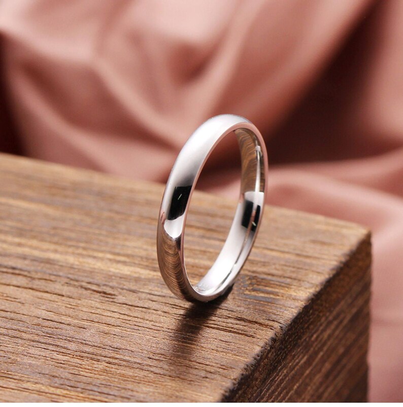 May include: A polished silver ring with a smooth, rounded band. The ring is set against a wooden surface with a soft pink background. The ring's reflective surface catches the light, highlighting its simple, elegant design. This ring is a piece of jewelry.