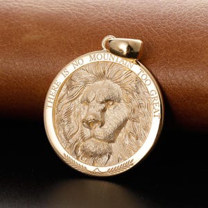 Könnte beinhalten: Goldfarbener Anhänger mit detailliertem Löwenkopf in der Mitte. Der Anhänger trägt die Inschrift "THERE IS NO MOUNTAIN TOO GREAT" um den Löwenkopf. Der runde Anhänger hat eine Schlaufe oben für eine Kette.