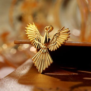Könnte beinhalten: Ein goldfarbener Anhänger in Form einer Taube mit ausgebreiteten Flügeln. Der Anhänger hat detaillierte Federn und eine Öse für eine Kette. Die Taube ist vor einem verschwommenen Hintergrund platziert, der das Design des Schmucks hervorhebt.