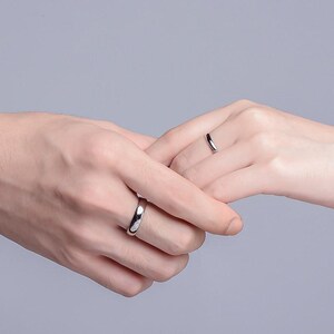 May include: Two silver rings, one on each hand, are shown in a close-up shot. The rings are simple bands with a polished finish. The hands are clasped together, suggesting a connection or commitment. The background is a soft, neutral gray.