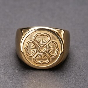 Può includere: Un anello a sigillo color oro con una faccia circolare caratterizzata da un design inciso a forma di fiore a quattro petali. Il centro del fiore presenta un piccolo cerchio con linee radianti che si estendono verso i petali, su una finitura metallica spazzolata.