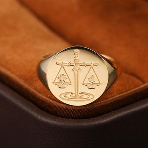 May include: A gold-toned signet ring featuring a circular face engraved with a central sword positioned vertically. The sword is flanked by a set of scales, with a sun symbol on the left balance pan and a crescent moon symbol on the right balance pan.