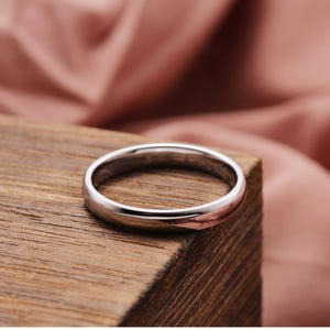 May include: A silver ring with a polished finish, resting on a wooden surface. The ring's smooth, rounded design reflects the light, showcasing its metallic sheen. The background features a soft, pink fabric.