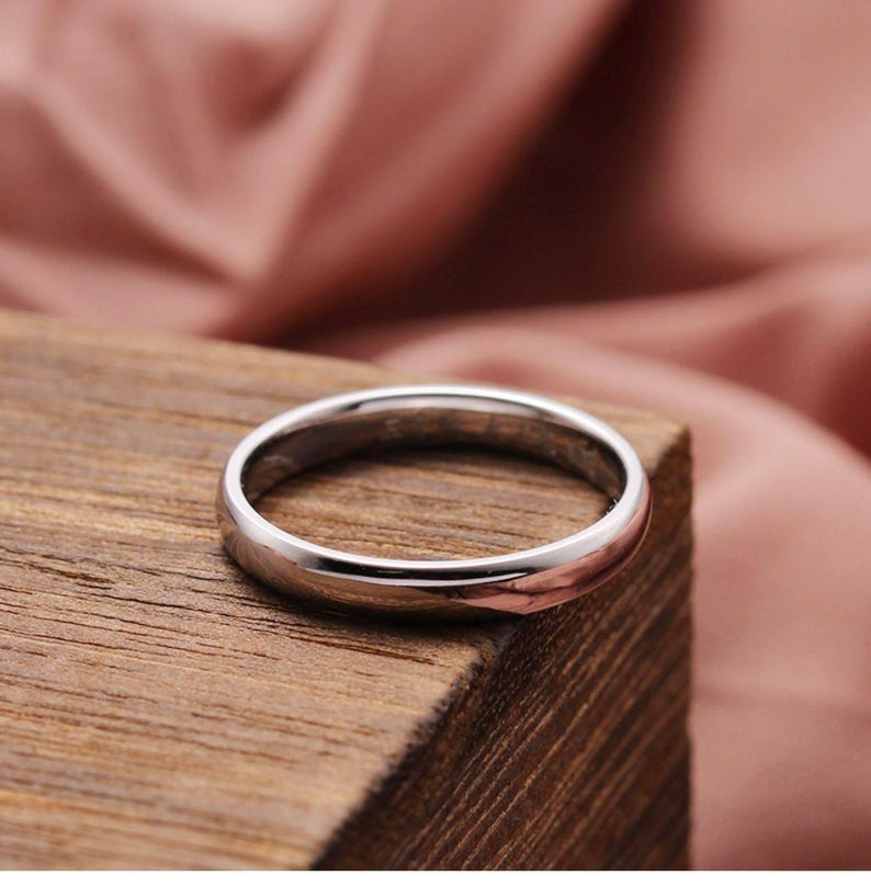 May include: A silver ring with a polished finish, resting on a wooden surface. The ring's smooth, rounded design reflects the light, showcasing its metallic sheen. The background features a soft, pink fabric.