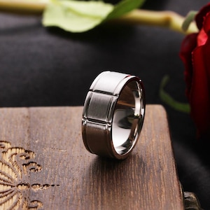 May include: A wide silver-toned metal ring featuring a brushed finish on the outer band. The surface of the ring is segmented by vertical grooves that create a series of rectangular panels around the circumference of the band.