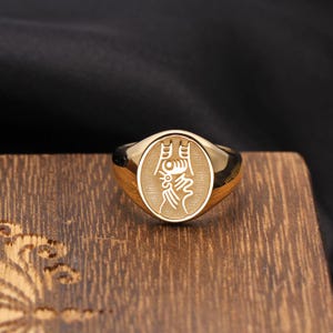 Puede incluir: Un anillo de sello dorado con una cara ovalada, fondo texturizado y un símbolo gráfico grabado en blanco. El anillo presenta una banda pulida y reflectante con una silueta suave y redondeada, mostrando un diseño de carácter detallado.