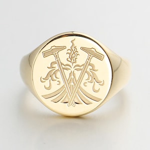 May include: A gold signet ring with a circular face. The face features a detailed engraving of two crossed hammers and ornate floral elements. The ring has a polished, reflective surface.