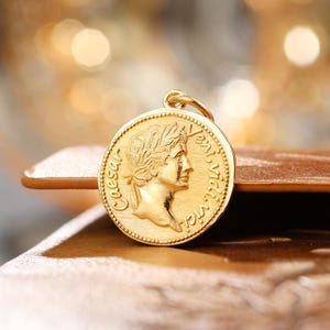 May include: A round gold pendant featuring a relief profile of Julius Caesar wearing a laurel wreath. The text Caesar and Veni, vidi, vici is embossed along the edge of the coin-shaped charm, which includes a small circular bail at the top.