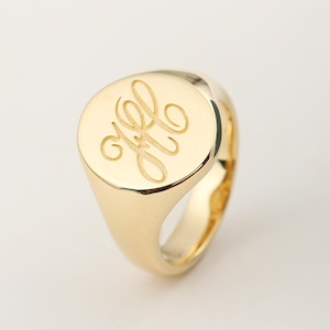 Könnte beinhalten: Ein goldener Siegelring mit poliertem Finish. Der Ring hat ein eingraviertes Monogramm auf der kreisförmigen Oberfläche. Das Band ist glatt und abgerundet. Der Ring wird vor weißem Hintergrund gezeigt.