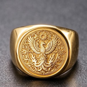 May include: A gold-toned signet ring featuring a circular face with an embossed phoenix design. The central bird is depicted with wings spread wide, surrounded by intricate scrollwork and a sun motif within a circular border on the ring face.