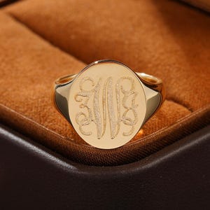 Pode incluir: Um anel de sinete dourado com uma face redonda polida e um monograma gravado com textura da letra W numa fonte cursiva decorativa. A banda é lisa e de ouro maciço, apresentando um design clássico com um acabamento metálico brilhante.