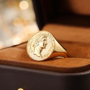 May include: A gold-colored signet ring featuring a detailed relief of a classical figure on a circular face. The ring is set against a brown background, showcasing its polished finish and historical design.