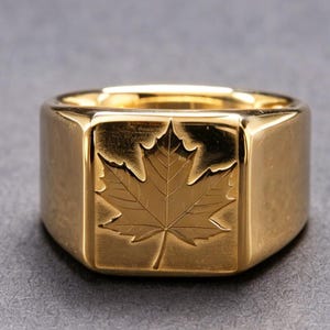 May include: A polished gold-tone signet ring featuring a square face engraved with a detailed maple leaf design. The ring has a smooth, reflective surface and a wide band, showcasing a clean, metallic finish throughout the entire piece.