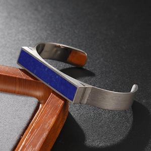 May include: A cuff bracelet featuring a rectangular, deep blue stone inlay set into a brushed, metallic silver-toned band. The bracelet has an open design with a rigid, geometric front section and a smooth, curved metal finish on the sides.