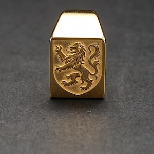 May include: A gold-toned metal ring featuring a rectangular face with a raised, detailed engraving of a rampant lion. The lion is depicted in profile, facing left, with its tail curled and paws raised. The ring has a polished, reflective finish.