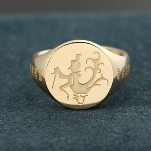 Puede incluir: Un anillo de sello dorado con un diseño grabado. El anillo presenta una cara circular con una ilustración detallada de una figura sosteniendo una espada. La banda del anillo tiene caracteres grabados en ambos lados. El anillo está sobre un fondo verde azulado oscuro.