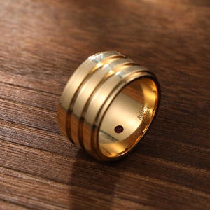 May include: A wide gold band ring featuring three parallel grooves along the exterior surface. The interior of the band is engraved with the text Au750 and 0.16ct, and it contains a small, circular, deep red gemstone set into the inner metal wall.