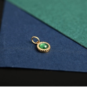 May include: A gold-colored pendant with a green oval-shaped stone. The stone is set in a textured, circular frame. The pendant has a small loop at the top for a chain. The background is a combination of blue and green.