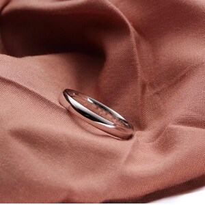 May include: A polished silver ring with a rounded band, resting on a textured, reddish-brown fabric. The ring reflects light, highlighting its smooth surface. The ring is a simple, elegant piece of jewelry.