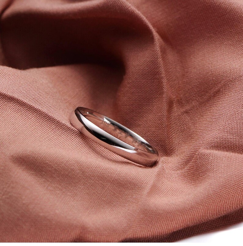 May include: A polished silver ring with a rounded band, resting on a textured, reddish-brown fabric. The ring reflects light, highlighting its smooth surface. The ring is a simple, elegant piece of jewelry.