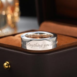May include: A polished silver-toned band ring featuring an engraved inscription that reads, "Anything you lose comes round in another form." The ring has a smooth, reflective surface and is displayed in a dark brown, velvet-lined jewellery box.