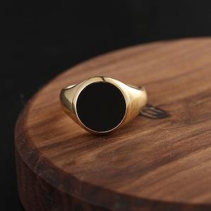 Puede incluir: Un anillo de sello dorado con una gran piedra negra circular. El anillo se muestra sobre una superficie de madera, destacando su acabado pulido y su diseño clásico. La banda del anillo tiene una forma lisa y redondeada.